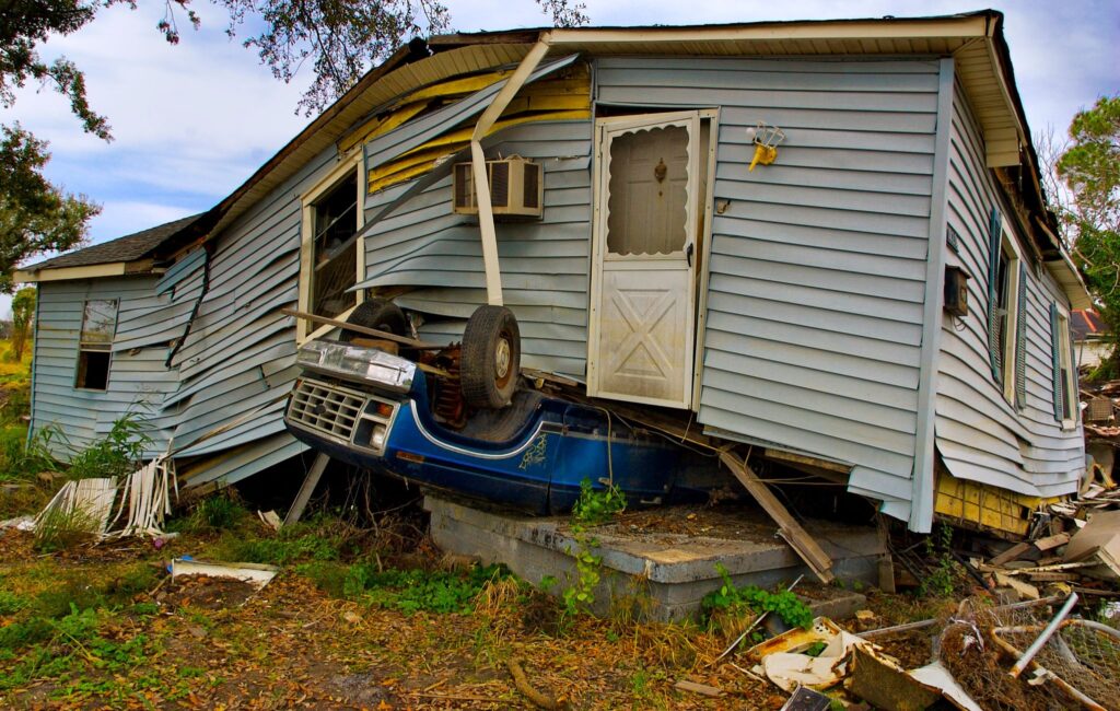 A mobile or manufactured home collapsing on top of a flipped-over car, surrounded by debris. This damage might’ve been prevented with a manufactured home foundation inspection.