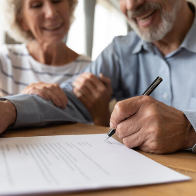 Zoomed-in image of elderly man and woman sitting together and signing a home inspection contract on paper, smiles on their faces.