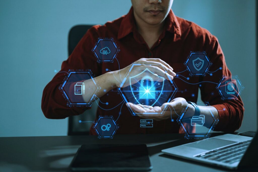 Man sitting with an open laptop and cell phone on the table in the foreground in front of him. He’s holding his hands in the air with digital hexagons drawn in the air featuring icons of a lock, phone, gears, a thumbprint, and other images of cybercrime and a cybersecurity insurance policy.