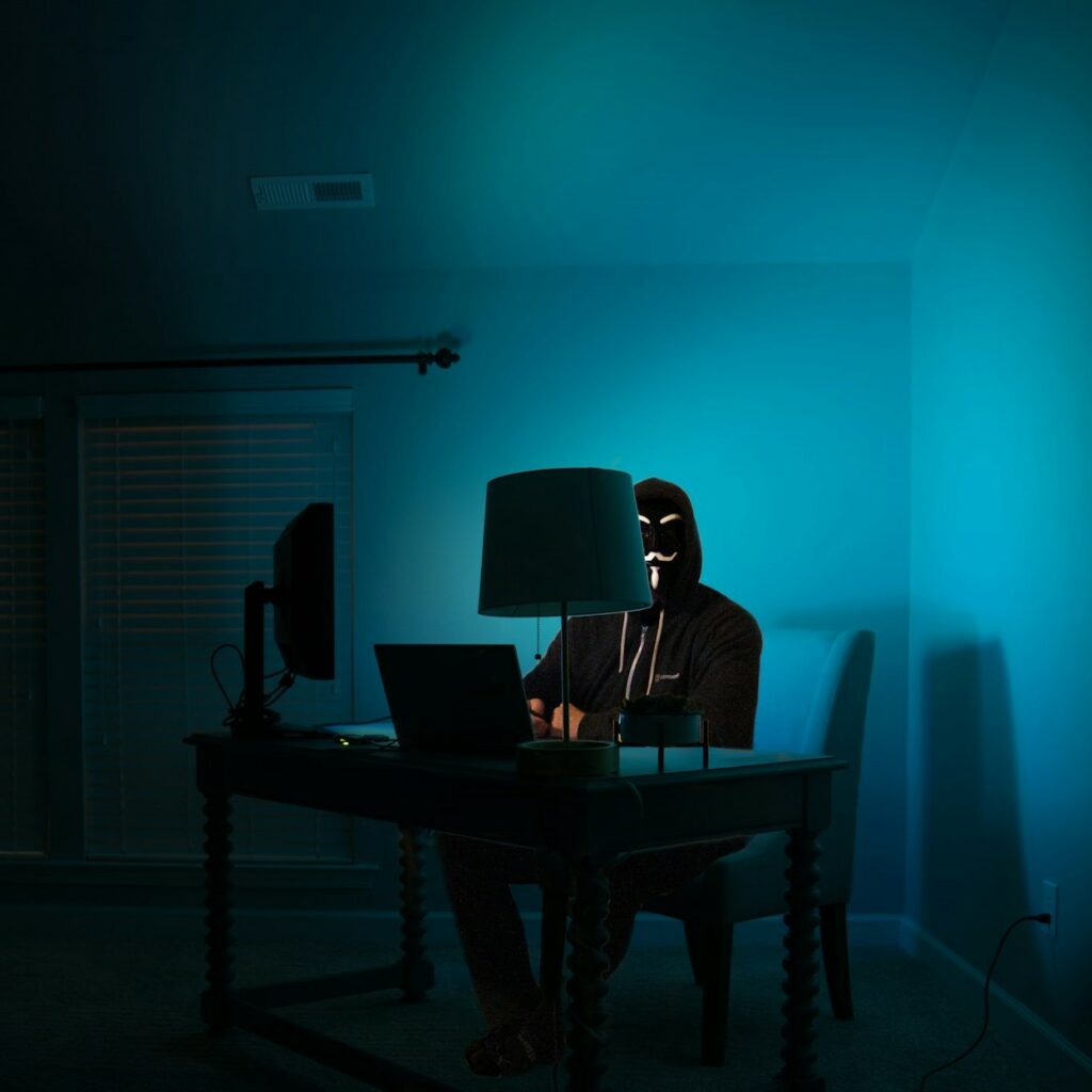 Person wearing an expressionless dark mask and black clothes sits in a dark room at a desk in front of a computer, illuminated only by a blue computer screen. Implies malicious ransomware or data theft.