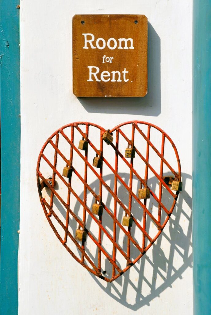 White and blue exterior wall with wooden sign reading "Room for Rent," above a hanging red, metal heart sign with small locks attacked to it. This rental space would raise accessibility concerns for a rental property inspection.