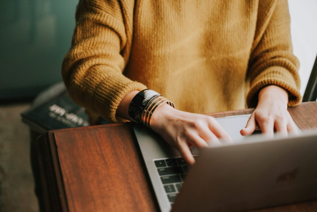 Person in mustard sweater using client communication skills to type effective communication with customers via email on laptop.