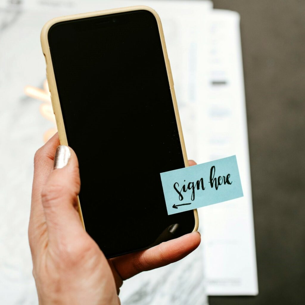 Close up of a hand holding a yellow smart phone with the screen turned off, and a blue sticky note pointing to the screen that reads "Sign here."