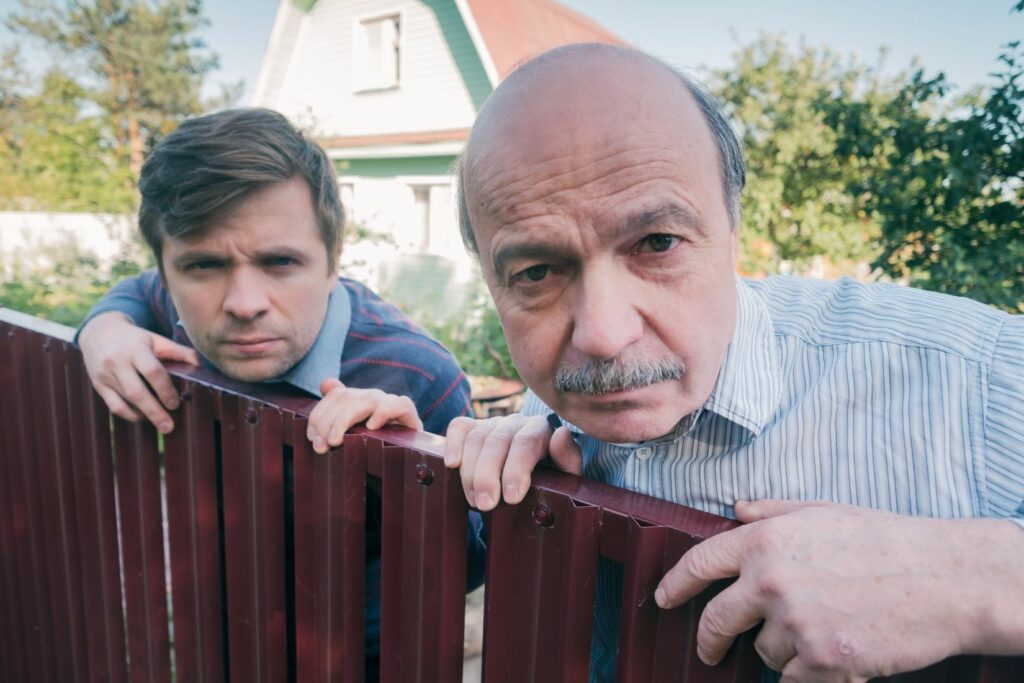 Two men with judgmental expressions leaning over a fence display the role of a nosy neighbor or interfering seller at a home inspection, as seen in this small claims court defense story.