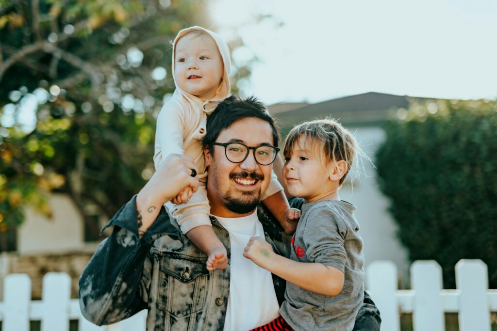 Man standing in front of white picket fence holds a toddler on his shoulders and another small child in his arms. Smiles at the camera. If you're wondering how much can you make as a home inspector, focus on the client's experience and make them happy, like this happy family.