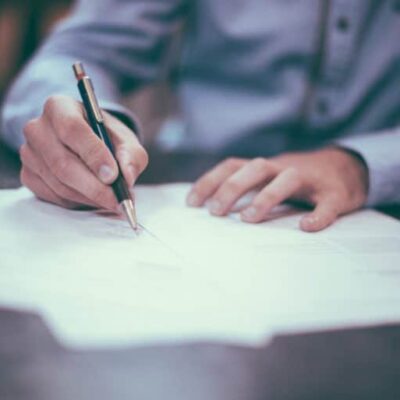 Zoomed-in image of a person’s hands writing on a stack of papers with a pen, as if signing someone’s home inspector pre-inspection agreement.