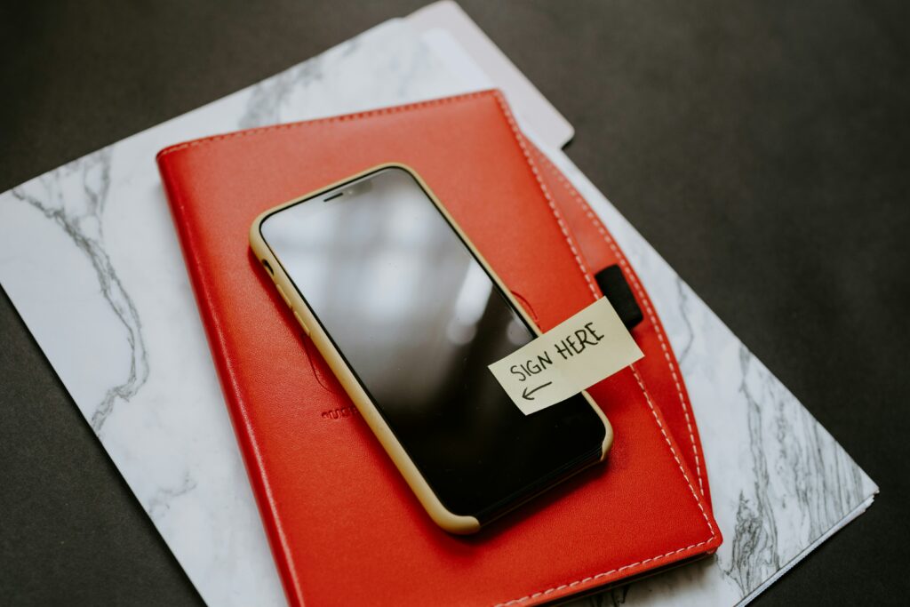 A table surface featuring a marbled paper folder, a red notebook, and a smartphone in a yellow case on top. On the phone is a sticky note that says "Sign Here." Represents a digital sewer scope inspection agreement that a client must sign ahead of time.