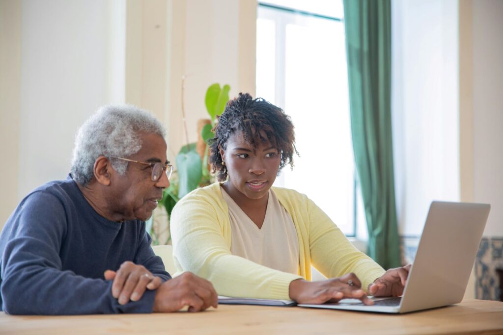 A woman looks at a laptop while typing. She sits next to an older gentleman, perhaps a home inspector mentor, who is also looking at the laptop screen.