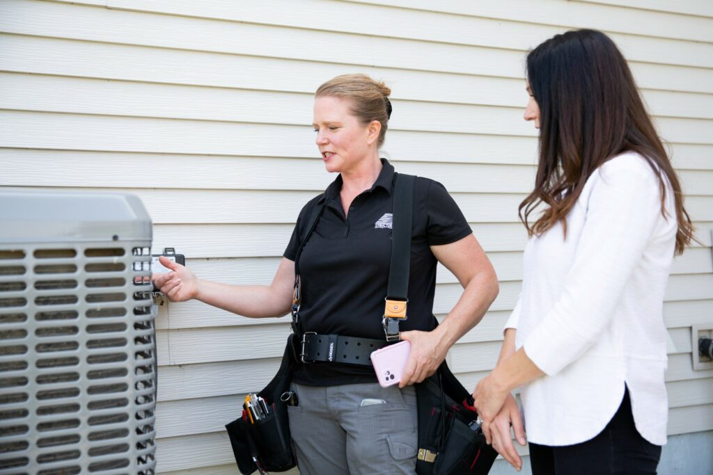 Person training to be a home inspector or learning from an inspector, together discussing the inspection next to an air conditioning unit. Photo courtesy of Structure Tech Home Inspections.