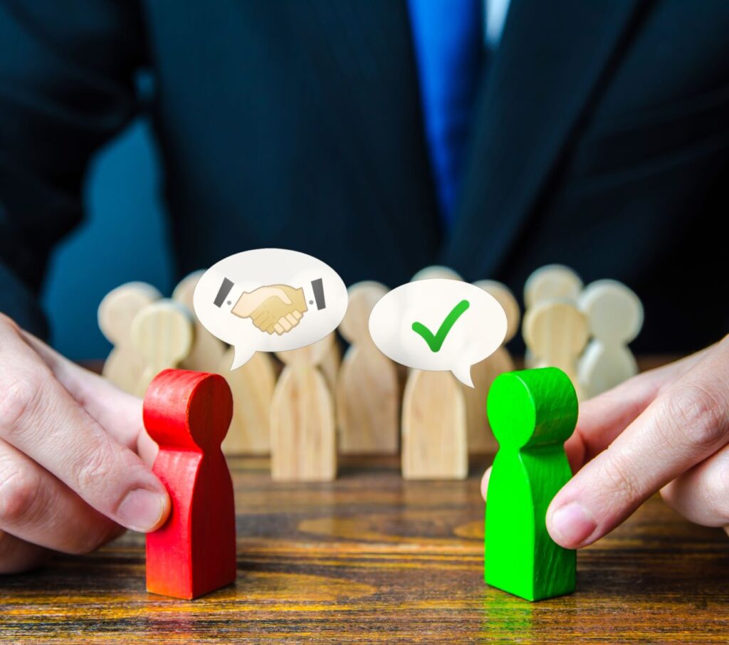 Person in business suit holding two wooden blocks shaped like people: a bright red one on the left with a "shaking hands" emoji above its head, and a green one to the right with a checkmark emoji above its head. Illustrates arbitration vs. mediation, where a middle party facilitates a solution to conflict.