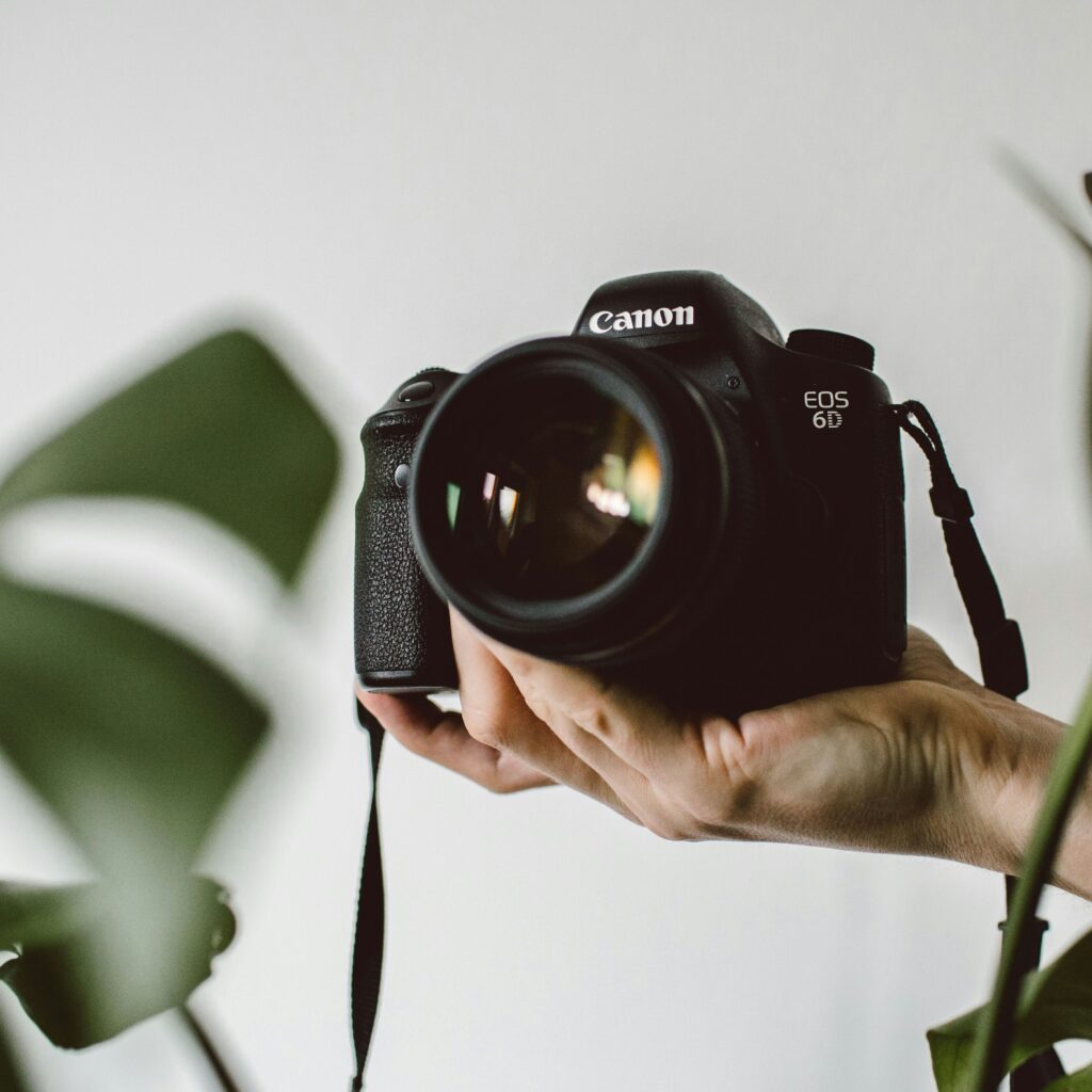 Person holds up a Canon camera with one hand, foliage in the background.