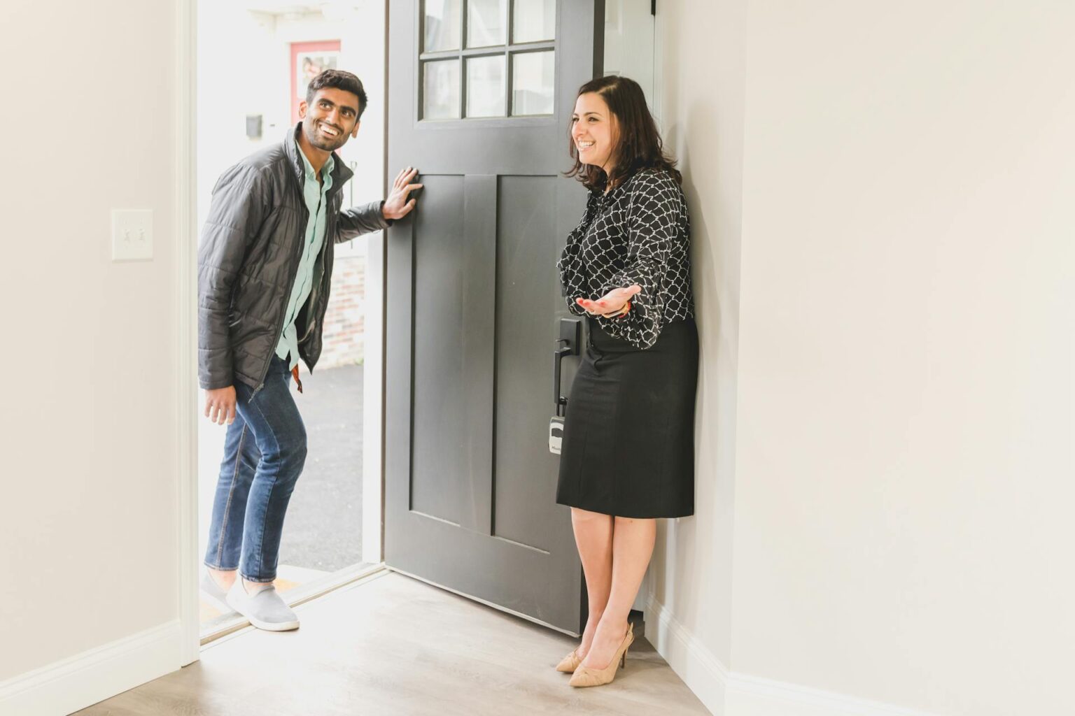 Woman stands holding a door open, smiling and gesturing into a house, while a man smiles and steps inside.