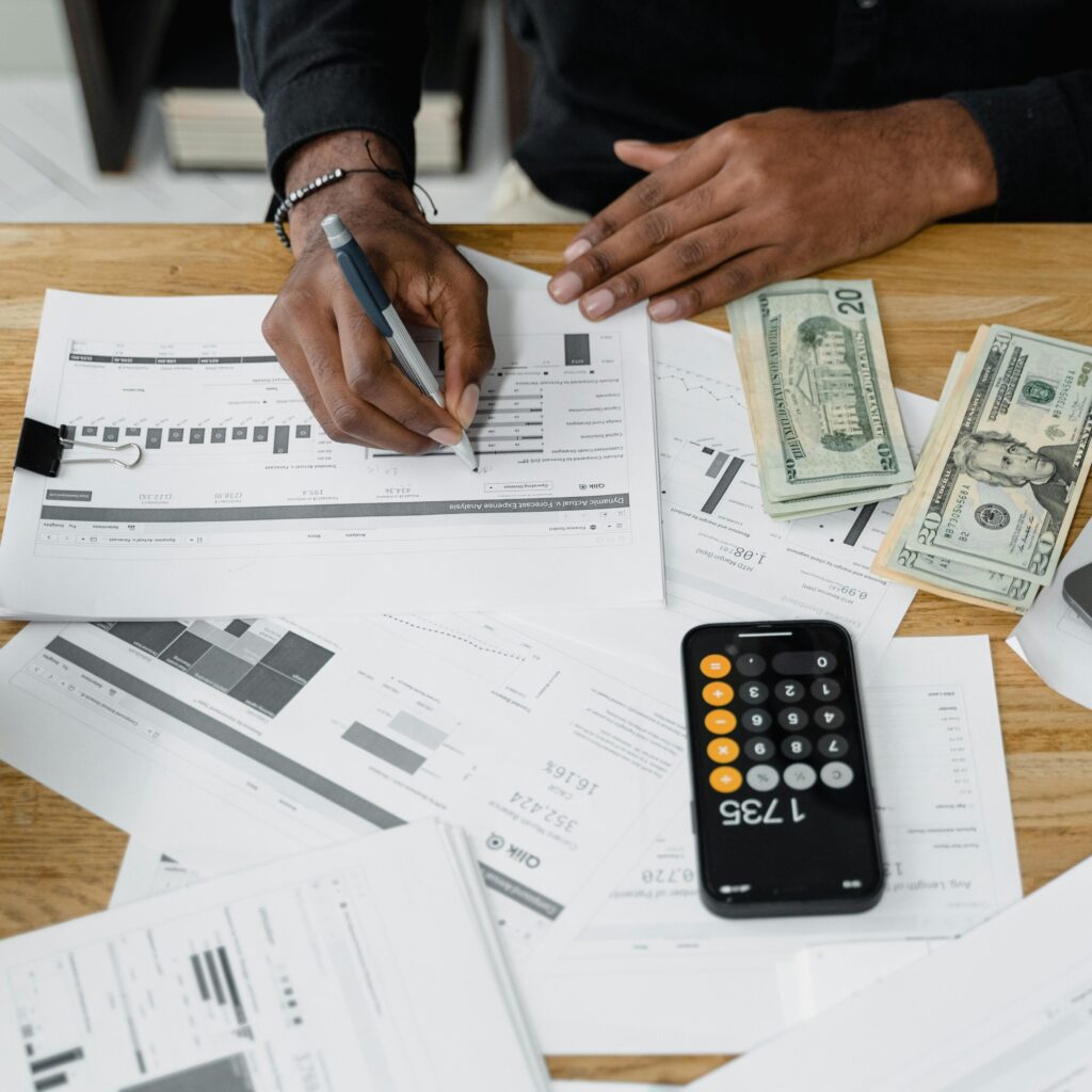 Person calculates numbers with a calculator while holding a pencil, with papers and cash scattered around.